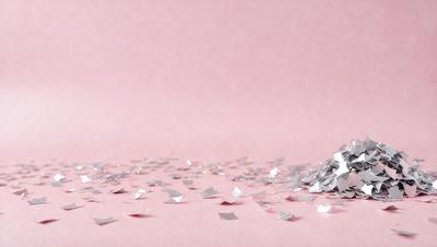 The primary subject of the image is a pink background with silver confetti scattered across it. The setting is a pink surface, and the visual style is a close-up of the confetti. The colors and mood of the image are predominantly pink and silver, creating a warm and festive atmosphere.
