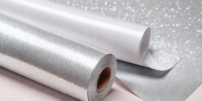 The primary subject of the image is a roll of silver metallic paper. The paper is shiny and reflective, giving it a metallic appearance. The setting is a table, and the visual style is a close-up of the paper roll. The colors and mood of the image are predominantly silver and reflective, creating a sense of sophistication and elegance.