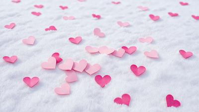 The primary subject of the image is a collection of pink hearts scattered across a white surface, which could be a snow-covered ground or a white sheet. The hearts are of various sizes and are placed all over the scene, creating a visually appealing and playful display. The setting appears to be a wintery environment, as the hearts are placed on a snow-covered ground. The visual style of the image is a combination of photo and digital art, as the hearts are arranged in a way that resembles a pattern