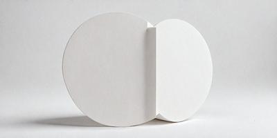 The primary subject of the image is a white cardboard box with a hole in the middle. The box is placed on a white surface, which serves as the setting and background of the scene. The visual style of the image is a photo, capturing the details of the cardboard box. The colors and mood of the image are predominantly white, giving it a clean and minimalist appearance.
