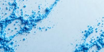 The primary subject of the image is a blue powder or substance, which is scattered across a white surface. The setting appears to be a laboratory or a clean room, as the background is predominantly white. The visual style of the image is a close-up of the blue powder, showcasing its texture and color. The dominant colors in the image are blue and white, creating a clean and professional atmosphere.