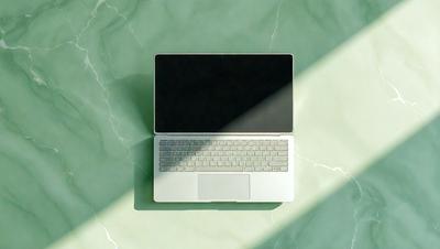The primary subject of the image is a white laptop computer. The laptop is placed on a green surface, which could be a table or a counter. The visual style of the image is a photo, capturing the laptop in detail. The colors in the image are predominantly white and green, giving the scene a clean and fresh appearance.