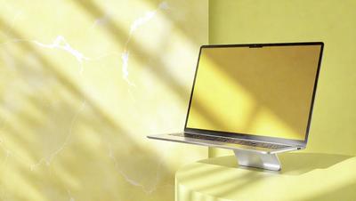 The primary subject of the image is a laptop computer. It is placed on a table, which is situated in front of a yellow wall. The visual style of the image is a photo, capturing the laptop and its surroundings. The colors and mood of the image are predominantly yellow, creating a warm and inviting atmosphere.