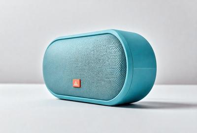 The primary subject of the image is a blue speaker, which is placed on a white surface. The speaker is a small, rectangular shape with a mesh-like design. The setting is a simple, clean environment, with no other objects or distractions visible in the image. The visual style is a close-up of the speaker, highlighting its unique design and color. The dominant colors in the image are blue and white, creating a calm and modern atmosphere.