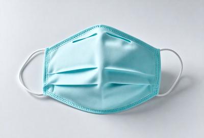 The primary subject of the image is a blue face mask. The mask is made of a soft, light blue fabric and features a white strap. The mask is placed on a white background, which provides a clean and uncluttered appearance. The visual style of the image is a close-up of the face mask, allowing the viewer to focus on its details. The colors and mood of the image are predominantly blue and calm, creating a sense of cleanliness and protection.