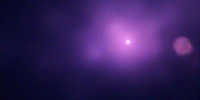 The primary subject of the image is a bright, glowing purple moon. The setting is a dark, cloudy sky, with the moon being the main focus of the scene. The visual style is a photo, capturing the moon's vibrant color and its position in the sky. The colors and mood of the image are predominantly purple, creating a serene and atmospheric ambiance.