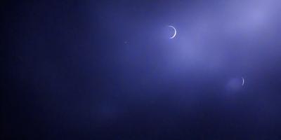 The primary subject of the image is the moon, which is seen in the sky. The setting is a dark, cloudy night with a blue sky. The visual style is a photo, capturing the moon's silhouette against the night sky. The colors and mood of the image are predominantly dark and moody, with the moon being the main source of light in the scene.