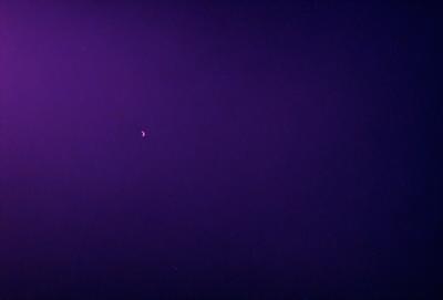 The primary subject of the image is a moon, which is located in the upper left corner of the purple sky. The setting is a dark, purple sky with no visible landmarks or objects. The visual style appears to be a photo, as it captures the moon and the sky in a natural, unaltered manner. The colors and mood of the image are predominantly purple, creating a serene and peaceful atmosphere.