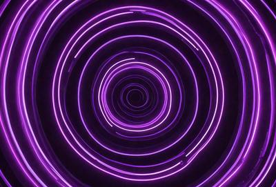 The primary subject of the image is a purple spiral, which appears to be a neon light or a purple circle. The setting is a dark background, which contrasts with the bright purple spiral. The visual style is a close-up of the spiral, emphasizing its shape and color. The colors and mood of the image are predominantly purple, creating a visually striking and somewhat mysterious atmosphere.