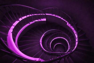 The primary subject of the image is a spiral staircase with a purple glow. The staircase is made of metal and has a purple light illuminating it. The setting is a dark room, and the visual style appears to be a combination of a photo and digital art. The colors and mood of the image are predominantly purple, creating a unique and atmospheric scene.