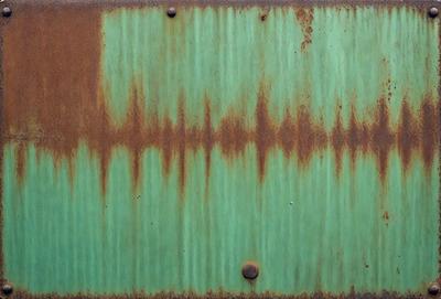 The primary subject of the image is a green metal surface with a rusted appearance. The setting is an outdoor environment, possibly a wall or a door. The visual style is a photo, capturing the rusted metal surface with its distinctive texture and color. The colors and mood of the image are predominantly green, with a hint of rust and a somewhat aged or weathered appearance.
