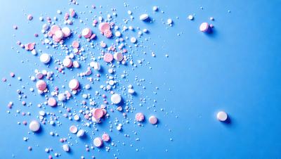 The primary subject of the image is a close-up of a blue surface with a large number of pink and white pills scattered across it. The setting appears to be a blue table or countertop. The visual style of the image is a close-up of the pills on the blue surface, creating a detailed and focused composition. The colors and mood of the image are predominantly blue and pink, giving it a calm and soothing atmosphere.