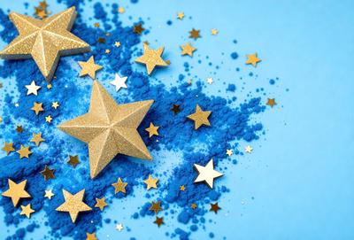 The primary subject of the image is a close-up of a star-shaped object, likely made of gold or gold-colored material, with blue stars surrounding it. The setting is a blue background, which complements the colors of the star-shaped object. The visual style appears to be a combination of a photo and digital art, as the star-shaped object is placed in a digital art-like environment. The colors and mood of the image are predominantly blue and gold, creating a warm and inviting