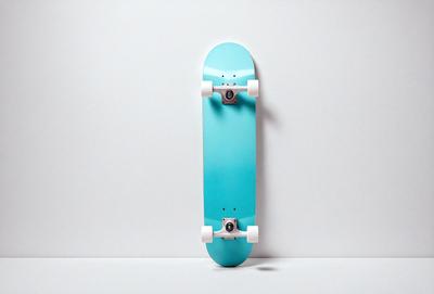 The primary subject of the image is a blue skateboard with white wheels. The skateboard is standing upright against a white wall, which serves as the setting and background of the image. The visual style appears to be a photo, capturing the skateboard in its natural environment. The dominant colors in the image are blue and white, creating a clean and modern atmosphere.