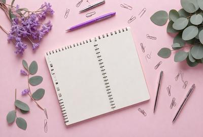 The primary subject of the image is a notebook with a pen and pencil on top of it. The setting is a pink surface, and the visual style is a photo. The colors and mood of the image are predominantly pink, creating a warm and inviting atmosphere.