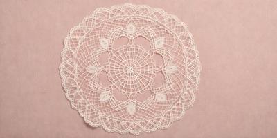 The primary subject of the image is a white lace doily with a pink background. The doily is a decorative piece that is typically used to protect the surface beneath it. The setting is a pink background, which adds a warm and inviting atmosphere to the scene. The visual style of the image is a close-up of the lace doily, showcasing its intricate details and texture. The colors and mood of the image are predominantly pink and white, creating a sense of elegance and