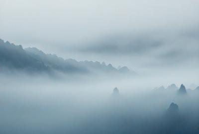 The primary subject of the image is a mountain range covered in fog. The setting is a mountainous area with a dense fog surrounding the peaks. The visual style is a photo of the mountain range, capturing the beauty of the foggy landscape. The colors and mood of the image are predominantly gray and moody, creating a sense of mystery and awe.