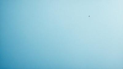 The primary subject of the image is a blue sky with a single black dot in the middle of it. The sky appears to be cloudless, and the black dot is the only visible object in the scene. The visual style of the image is a simple, minimalist representation of the sky. The colors and mood of the image are predominantly blue, creating a calm and serene atmosphere.