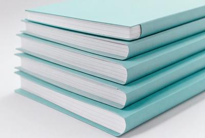 The primary subject of the image is a stack of books. The books are stacked on top of each other, and they are all of the same color, which is a shade of blue. The setting is a white surface, likely a table, where the books are placed. The visual style of the image is a close-up of the stack of books, showcasing their color and texture. The colors and mood of the image are calm and soothing, as the blue color of the books creates a sense of tranquility