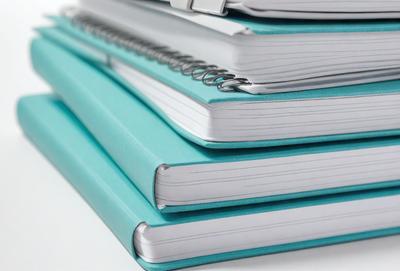 The primary subject of the image is a stack of spiral-bound notebooks. The notebooks are blue and have a silver spiral binding. The setting is a table, and the visual style is a photo. The dominant colors are blue and silver, creating a calm and organized atmosphere.