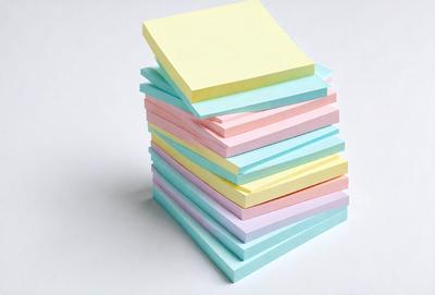 The primary subject of the image is a stack of colorful notepads. The setting is a white background, and the visual style appears to be a photo. The colors and mood of the image are vibrant and lively, with the various colors of the notepads creating a visually appealing and cheerful atmosphere.