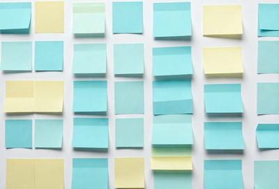 The primary subject of the image is a collection of blue and yellow sticky notes. These notes are arranged in a visually appealing manner, with some of them overlapping each other. The setting is a white surface, which provides a clean and uncluttered backdrop for the sticky notes. The visual style of the image is a combination of photo and digital art, as the sticky notes appear to be digitally manipulated to create a cohesive and organized arrangement. The colors and mood of the image are pre