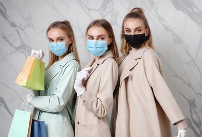 The image features three women wearing face masks and gloves, standing together in a room. They are all dressed in coats, with one woman wearing a white coat and the other two wearing tan coats. The women are positioned in a line, with one woman on the left, another in the middle, and the third on the right. They are all holding shopping bags, indicating that they might be shopping together or have recently completed a shopping trip. The overall atmosphere of the image is one of unity