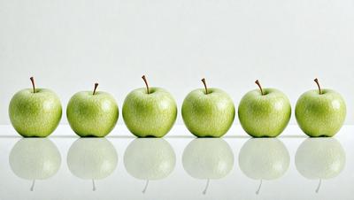 The primary subject of the image is a row of five green apples. They are arranged in a straight line, with each apple being slightly larger than the one next to it. The setting is a white background, which provides a clean and minimalist appearance to the image. The visual style is a close-up of the apples, emphasizing their details and texture. The colors and mood of the image are predominantly green, with a slightly cool and fresh atmosphere.