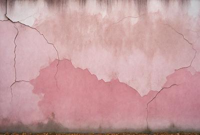 The primary subject of the image is a pink wall with a cracked and peeling surface. The setting is an outdoor environment, with the wall being part of a building. The visual style is a photo, capturing the pink wall and its damaged appearance. The colors and mood of the image are predominantly pink, giving it a warm and somewhat distressed atmosphere.