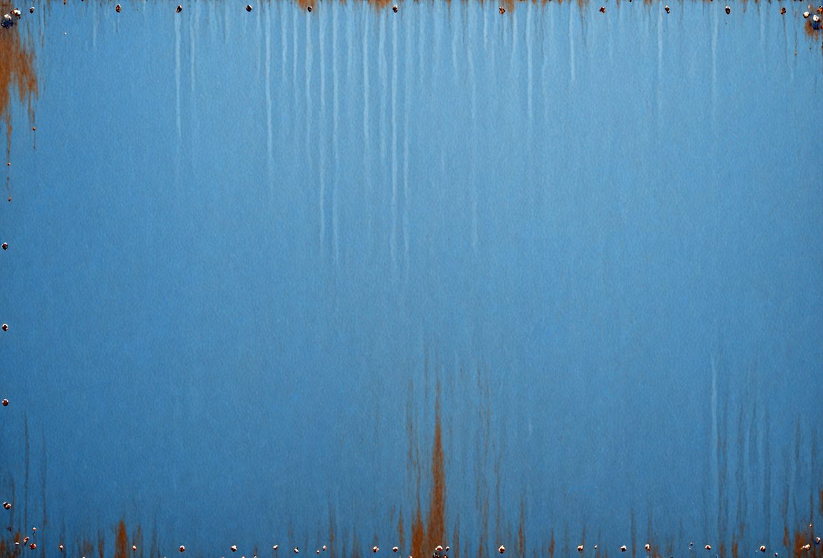 The primary subject of the image is a blue metal surface with rust stains. The setting is an outdoor environment, and the visual style appears to be a photo. The dominant colors are blue and rust, creating a mood of aged and weathered material.