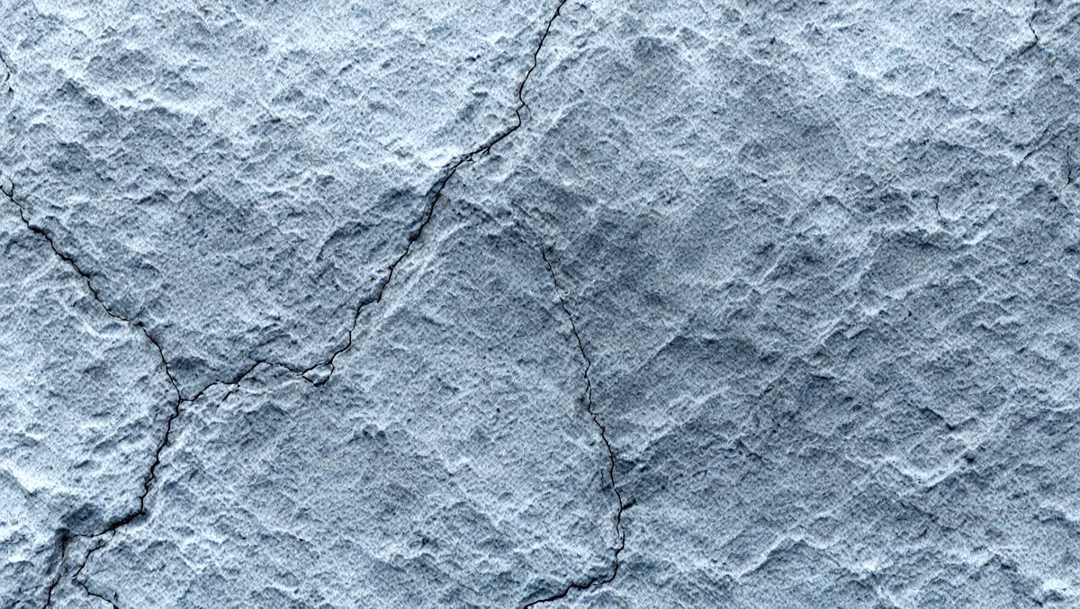 The primary subject of the image is a cracked, textured surface, possibly a stone wall or a concrete wall. The setting is an outdoor environment, with the cracked surface as the main object. The visual style is a close-up of the cracked surface, which gives a sense of depth and texture. The colors and mood of the image are predominantly gray and blue, with the cracked surface adding a sense of age and wear.